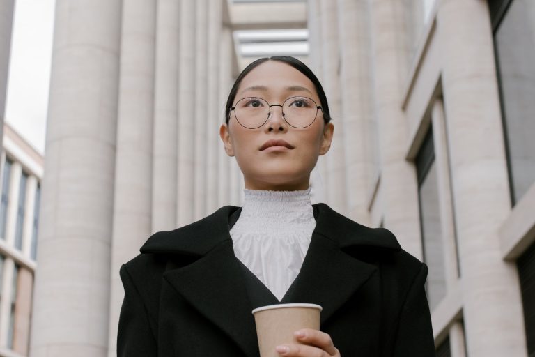Low-angle view of a composed woman in a black coat holding a coffee cup among stone columns
